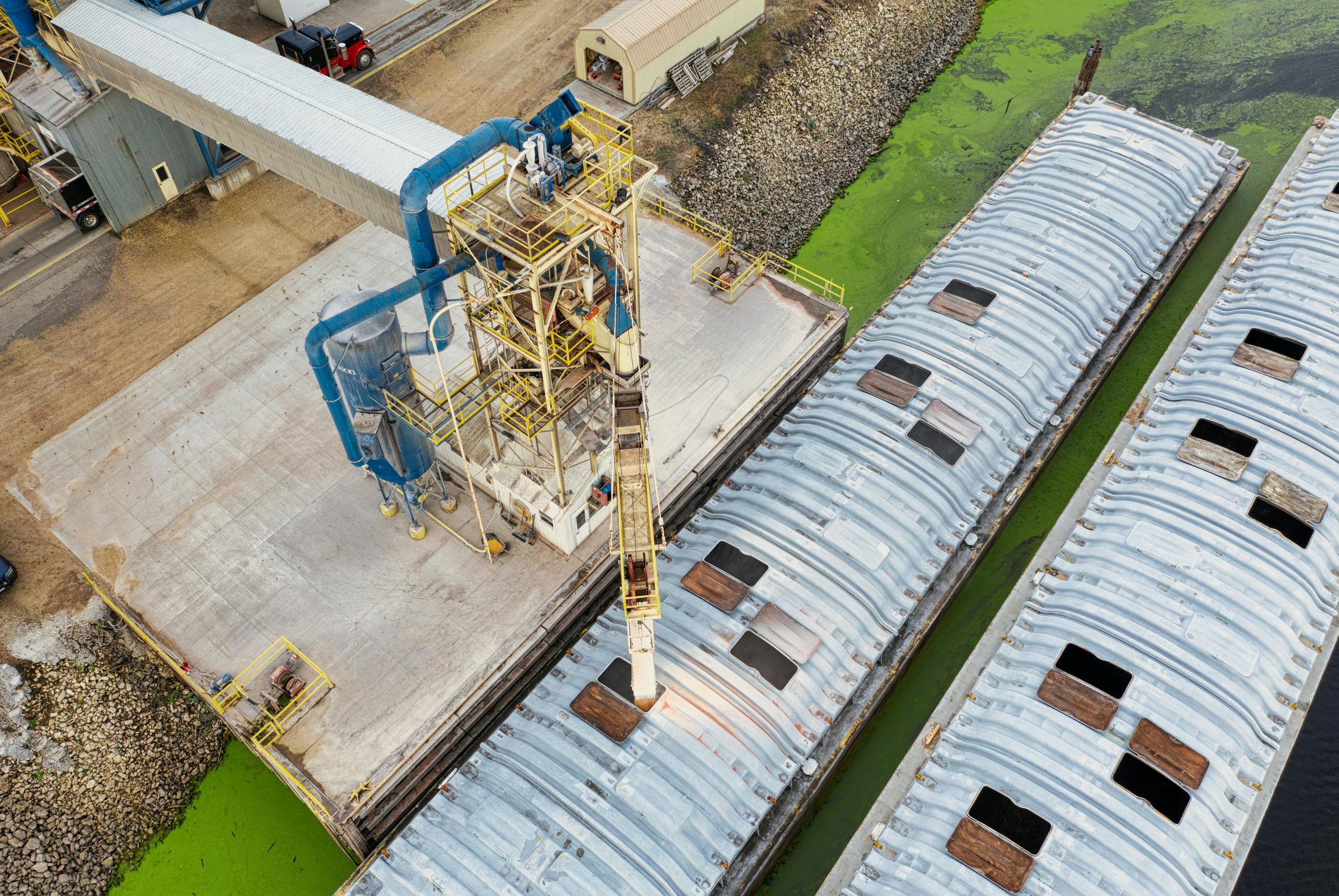 Aerial view of industrial barges and docking facility near green algae-infested water.